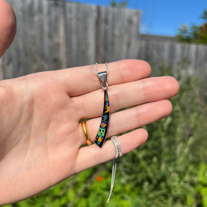 Hand holding men's elongated Southwest-style opal inlay pendant in sunlight against blurred outdoor background.