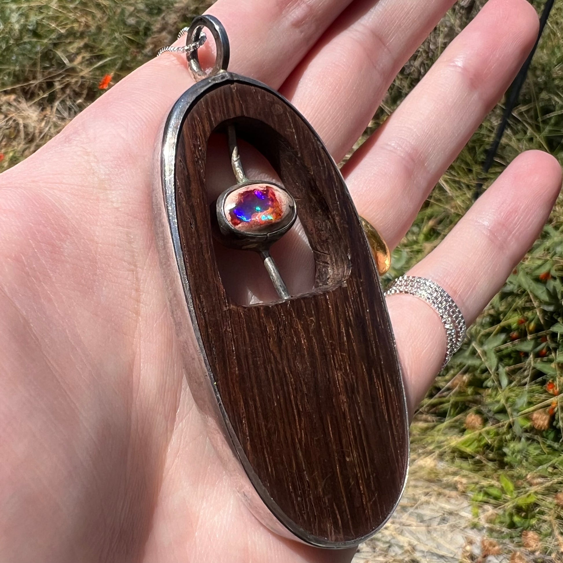 Hand holding a men's Mexican fire opal necklace with a carved, polished wood design in sterling silver in the sunlight.  Viewed from an angle, the opal has purple and green flashes.