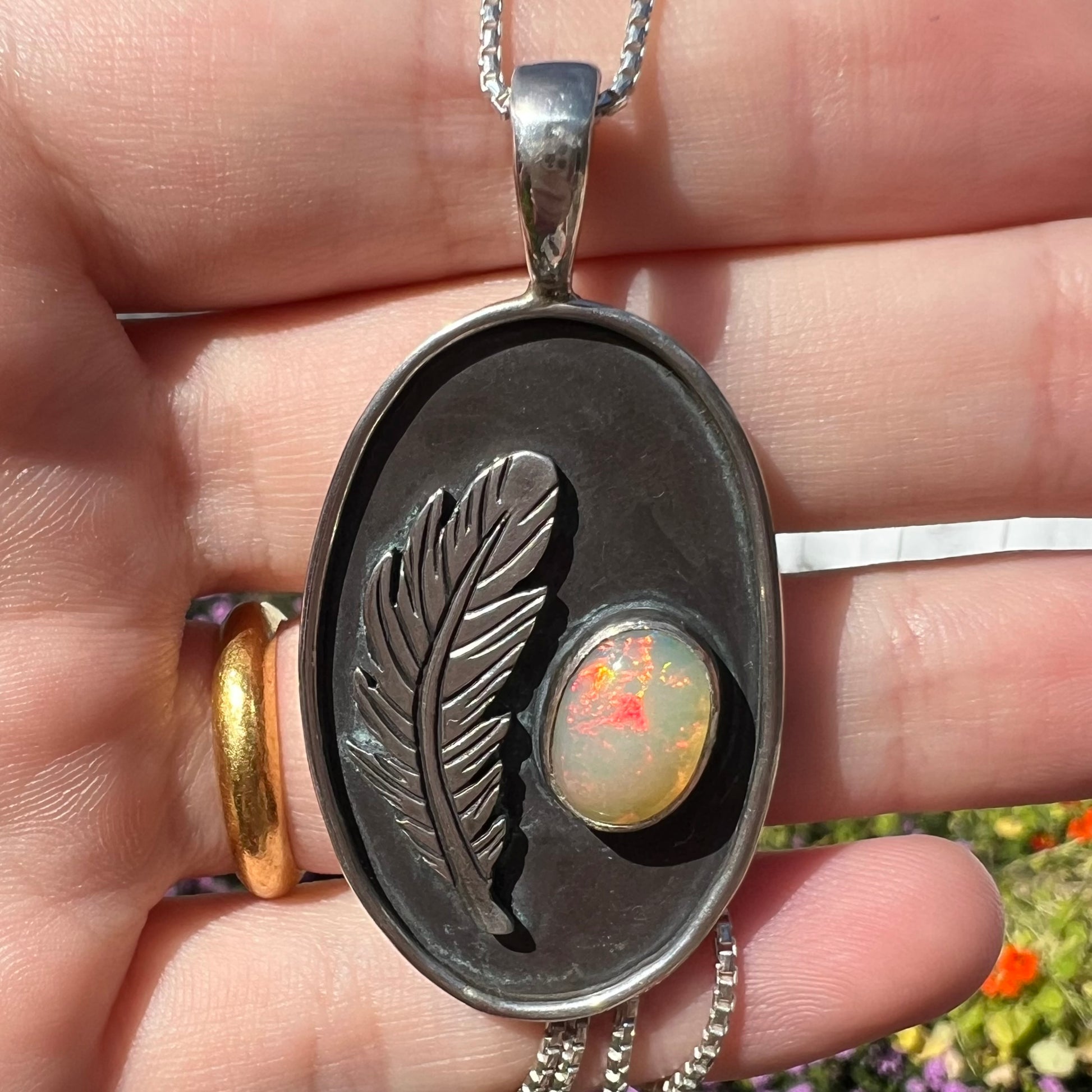 Close-up macro shot of men's fire opal and feather design necklace held in a hand under natural light.  The opal has red play of color.