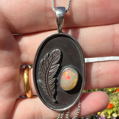 Close-up macro shot of men's fire opal and feather design necklace held in a hand under natural light.  The opal has red play of color.