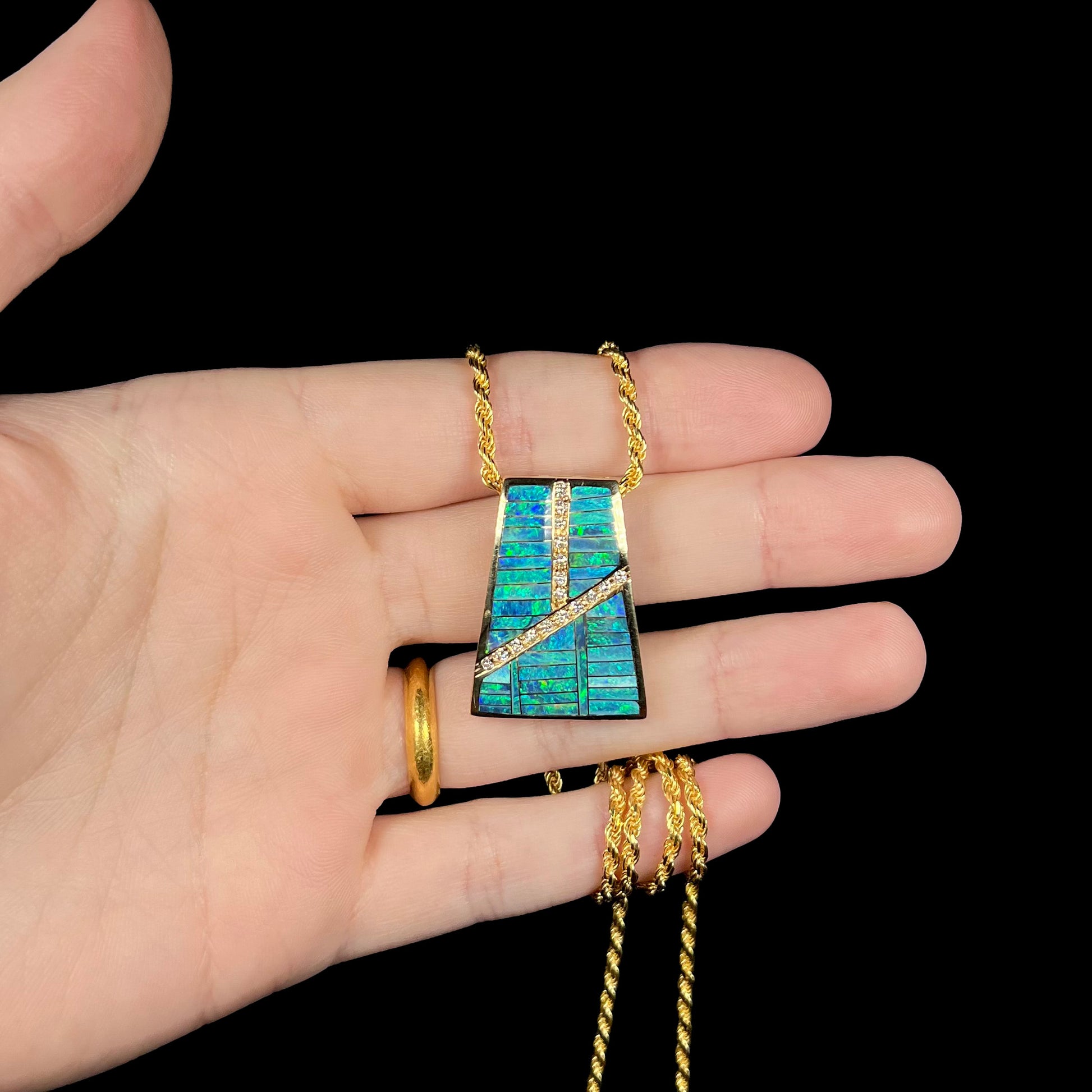 Hand holding a men's solid gold opal inlay and diamond necklace against a black background.