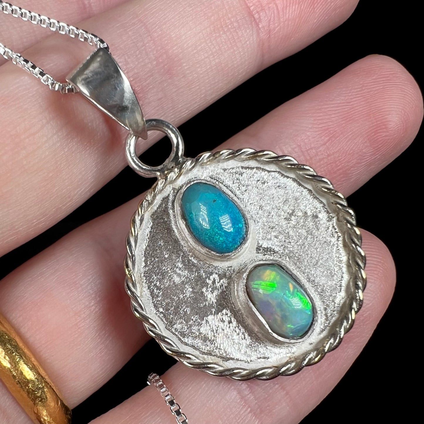Angled close-up of a men's handmade sterling silver Australian opal two-stone pendant with a rope bezel accent, resting against fingers.