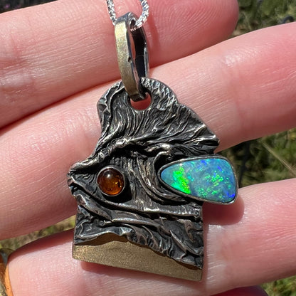 Close-up of a men's handmade sterling silver and 18k gold opal doublet and amber pendant held in the sunlight.  The opal has green flashes.