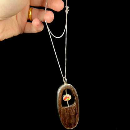 Hand dangling a men's handmade sterling silver fire opal and wood necklace from its chain.  The opal has a green flash.