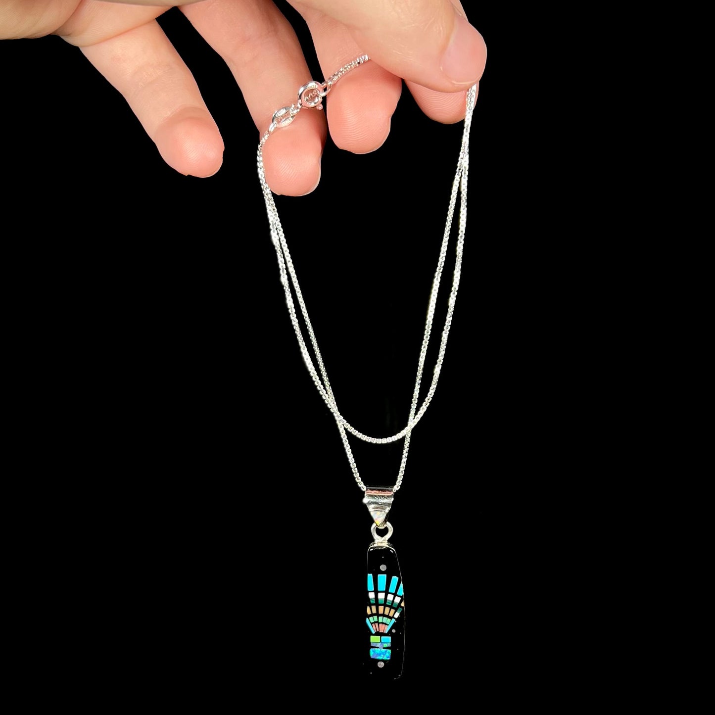 Hand dangling a sterling silver stone inlay necklace from its box chain in front of black background.