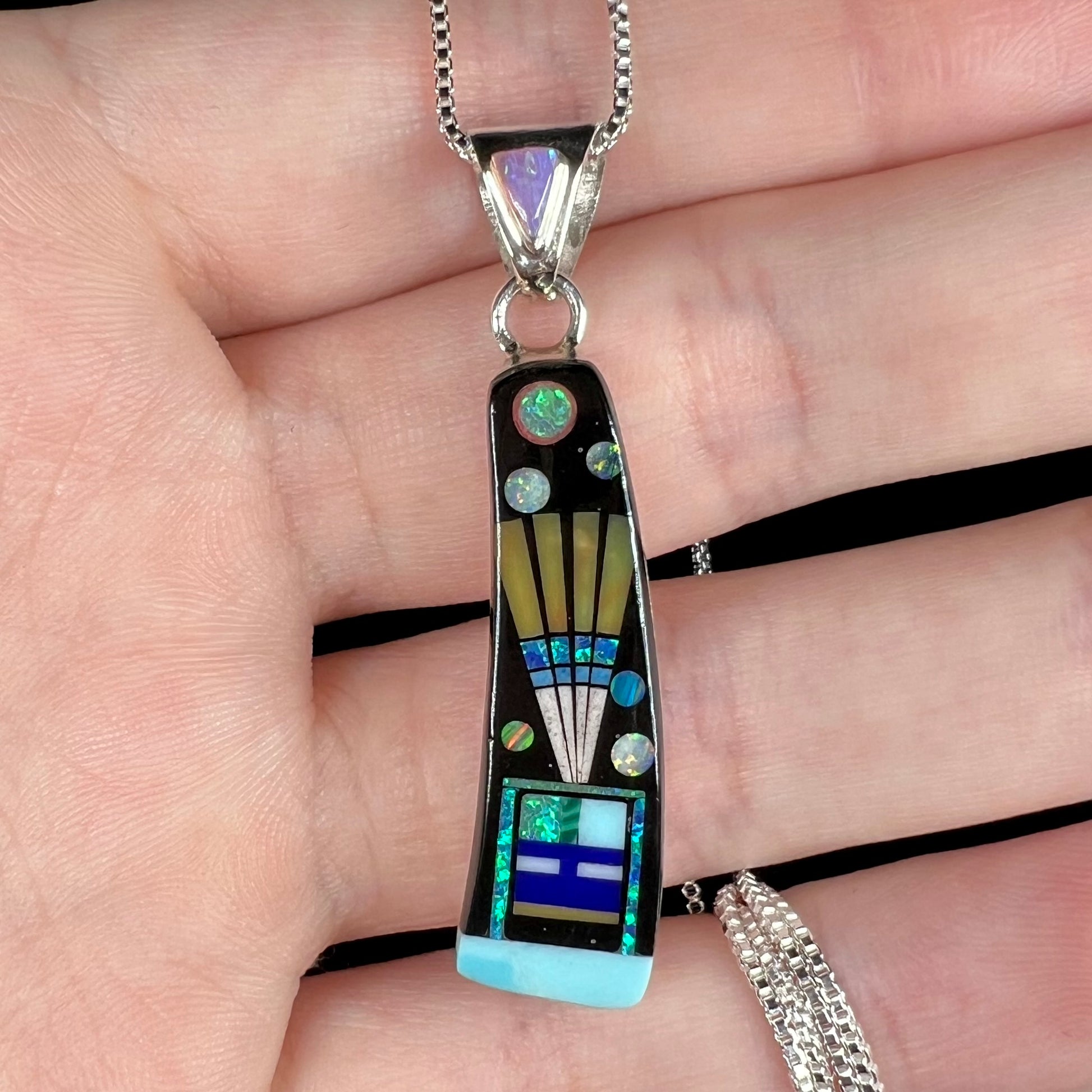 Close-up of men's inlay pendant with kachina and space motif held in hand under studio light.