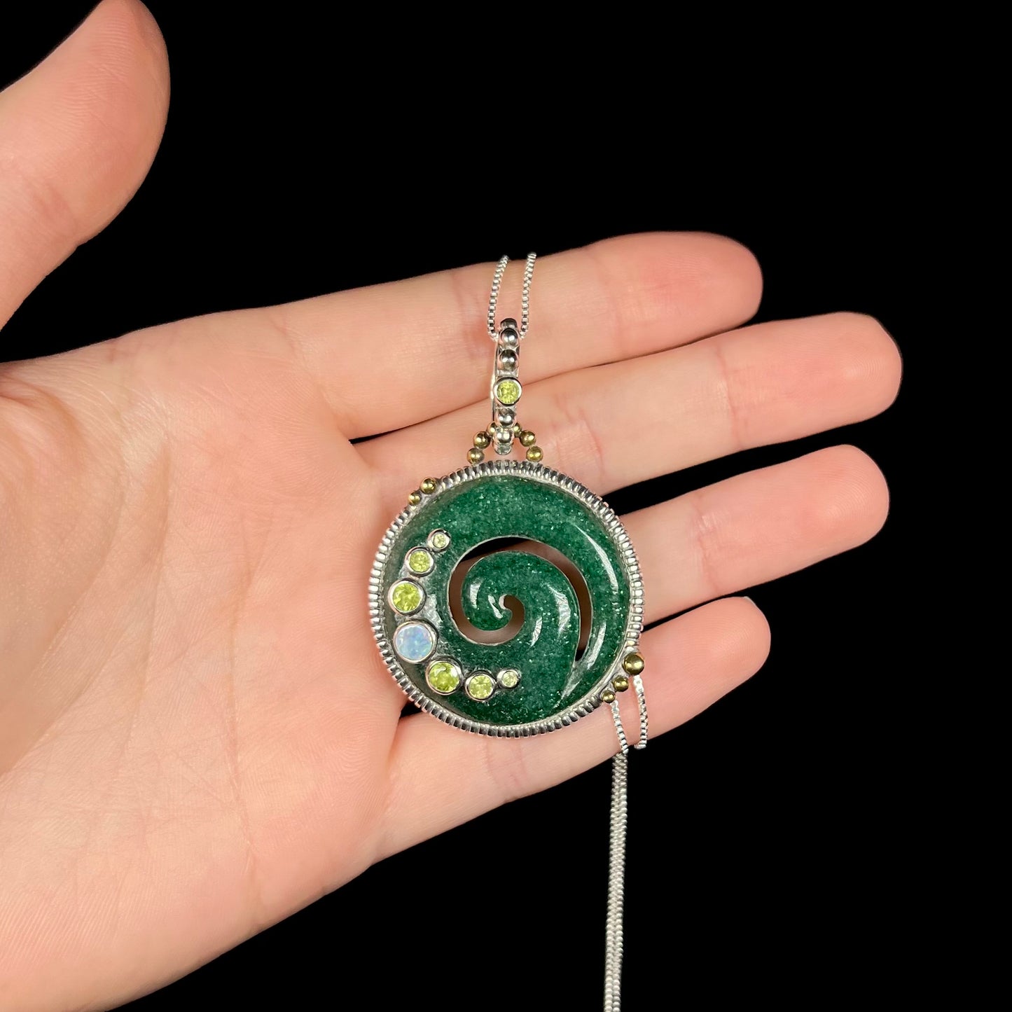 A men's aventurine koru necklace, accented with opal and peridot, shown in a hand against a black background.