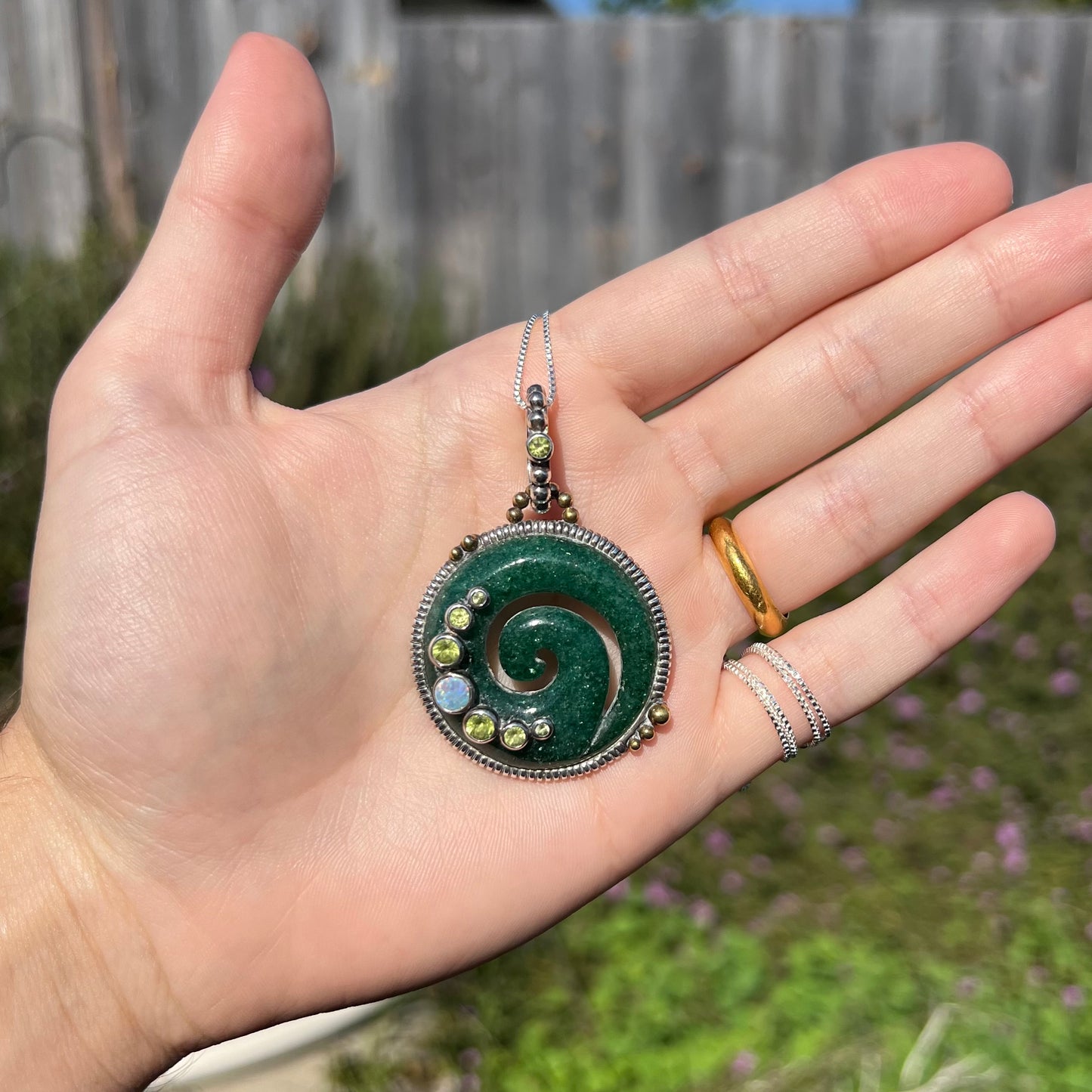 A men's aventurine koru necklace, accented with opal and peridot, shown in a hand in sunlight.