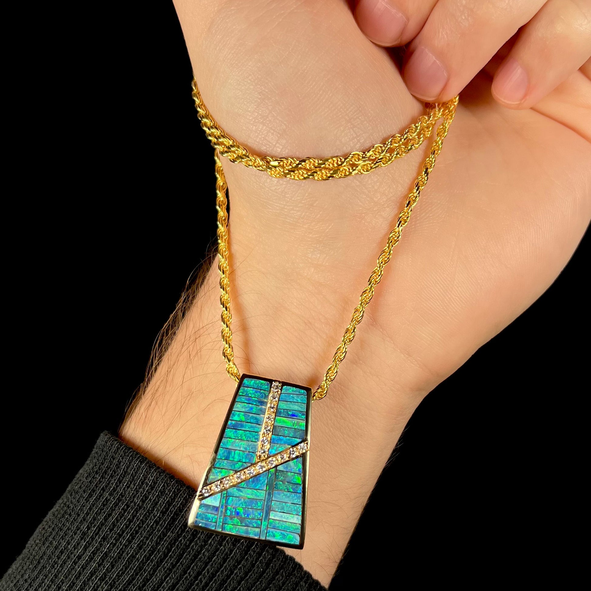 A hand holding a men's gold opal and diamond necklace by its rope chain.  The pendant is dangling at an angle against the person's wrist.