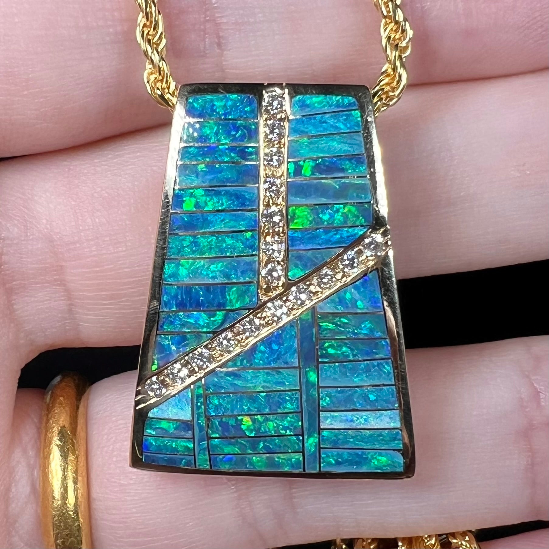 Close-up of a men's opal and diamond pendant held in a hand in cool LED lighting.  The opals appear blue and green.