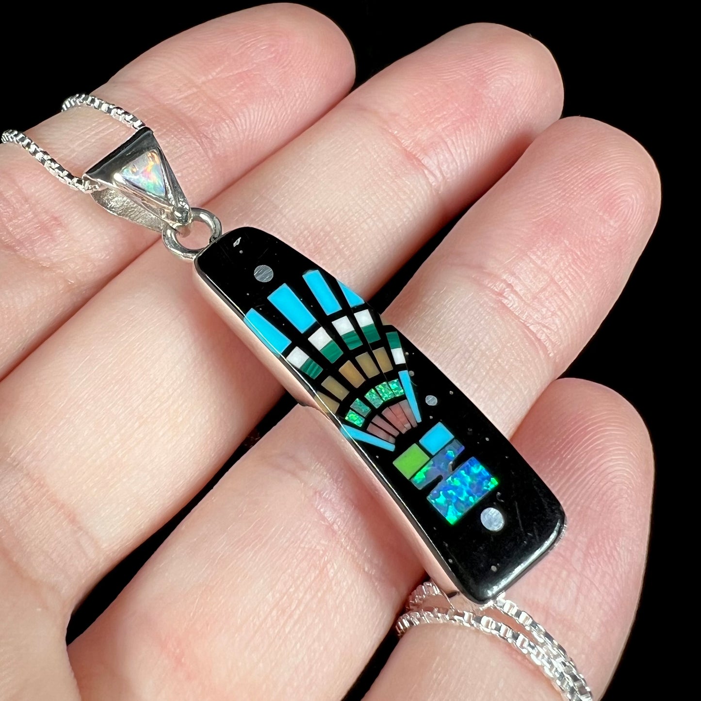 Angled close-up of men's opal and onyx inlay pendant with Southwest-style motif, held in hand against black background under cool studio light.