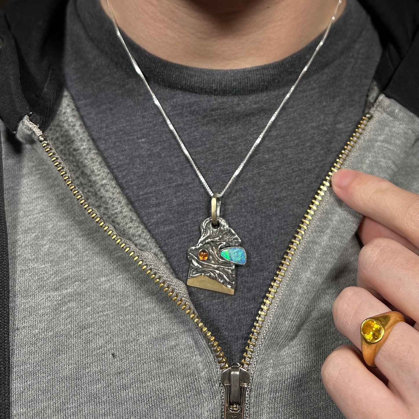 Close-up of a person wearing a men's opal and amber pendant in sterling silver by Starborn Creations against a dark shirt beneath a hoodie.