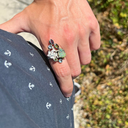 A silver men's ring set with rough Virgin Valley opal and gems with a female figure design, worn on a hand resting on a hip.