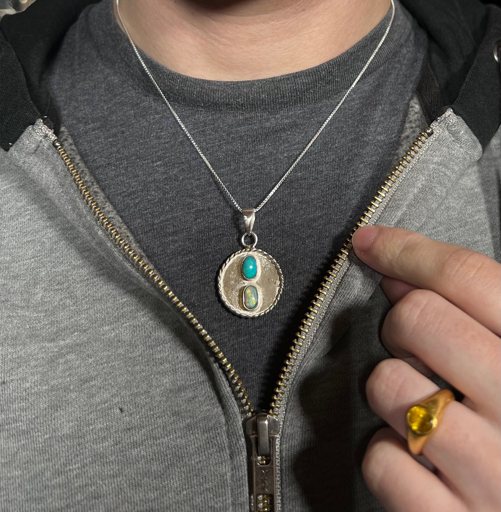 A men's sterling silver two-stone Australian opal medallion necklace worn around a person's neck against a dark shirt under an unzipped hoodie.