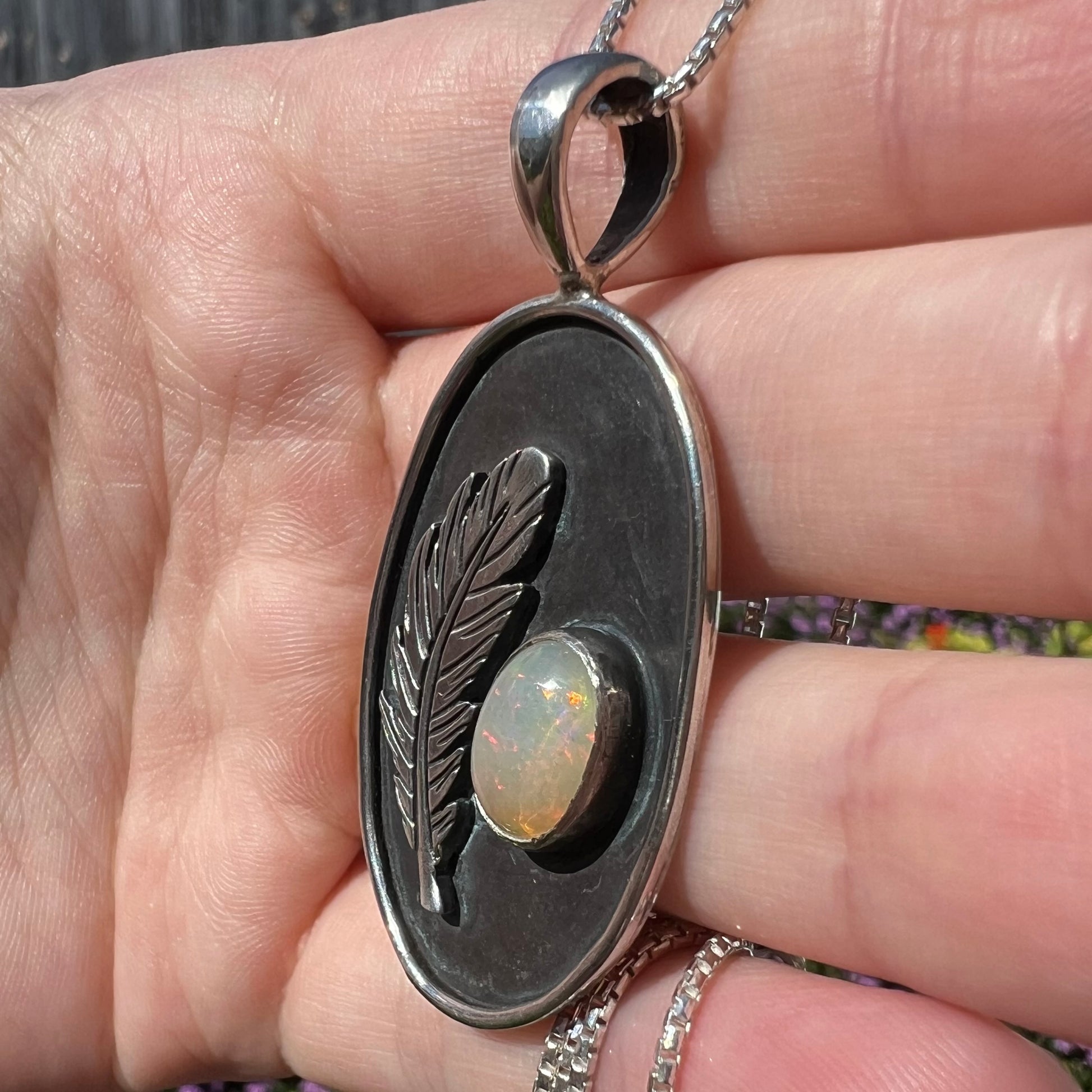 Close-up side view of a men's silver Ethiopian opal and feather design pendant held in a hand, showing the piece in sunlight.