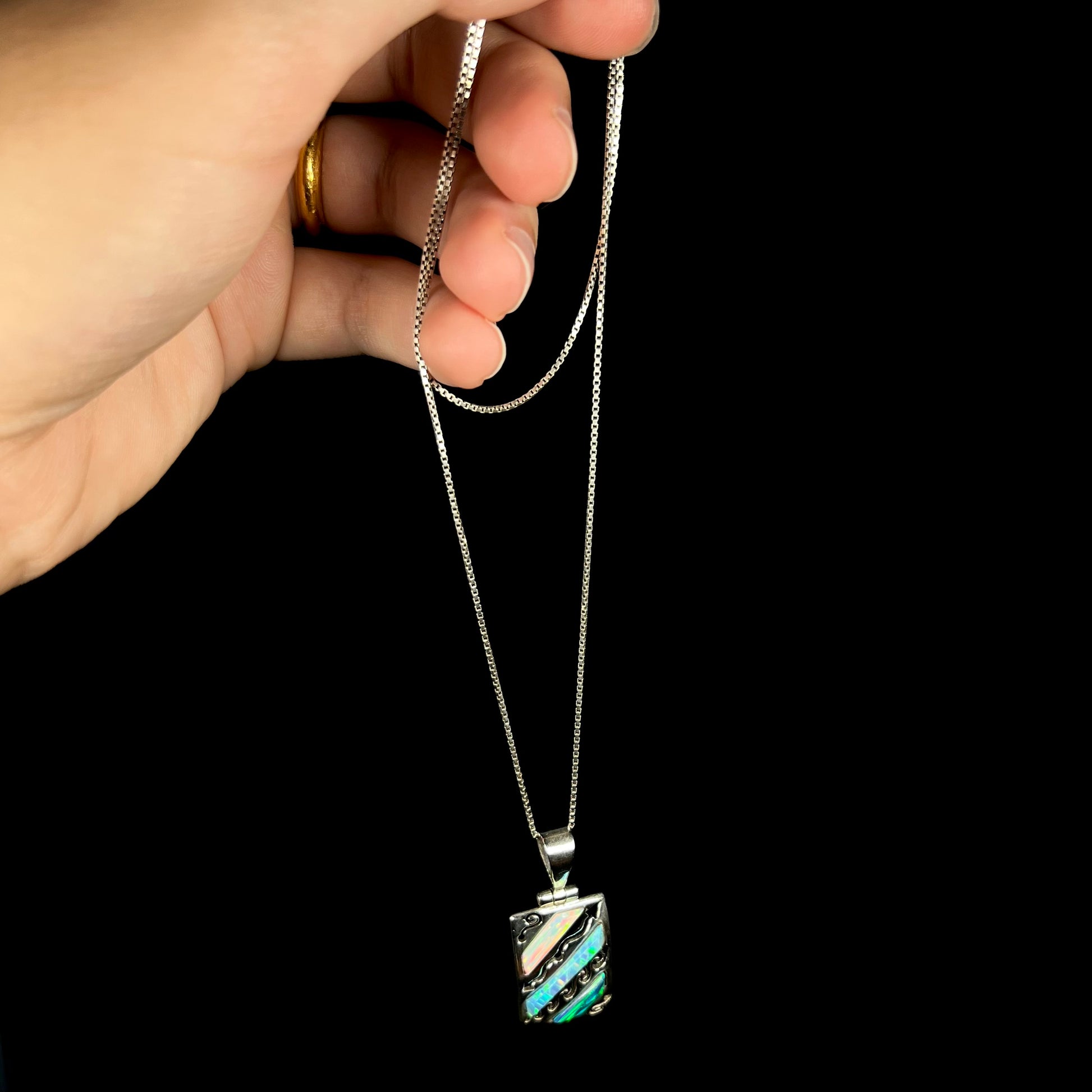 A men's 950 silver lab-created opal necklace, dangled from a hand by its chain against a black background.