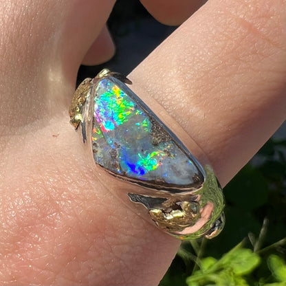 Angled close-up of a finger wearing a men's sterling silver and gold nugget opal ring in the sunlight.  The opal has rainbow flashes of color.