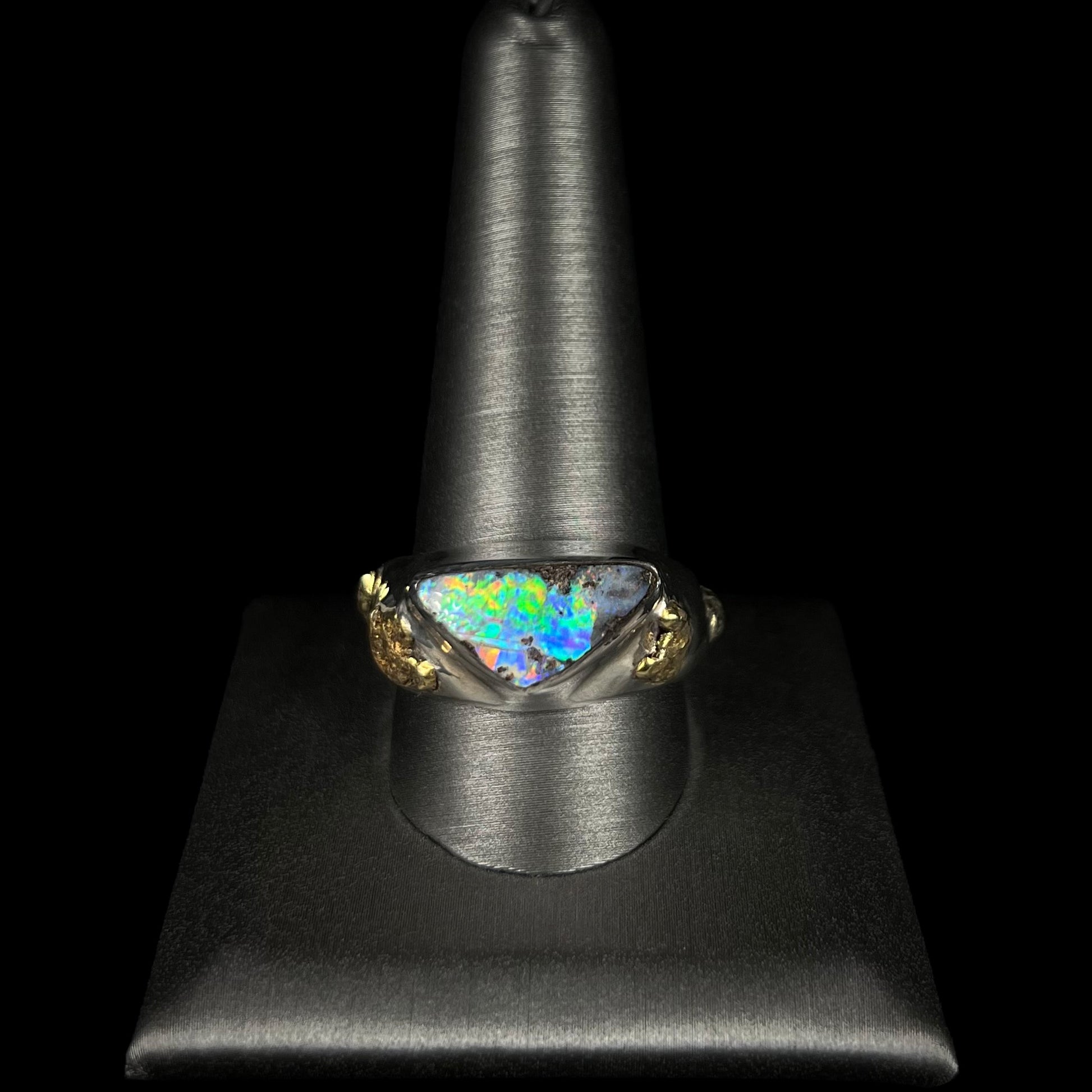 Distant front view of a men's size 12 sterling silver opal and gold nugget ring on a ring display.  The opal is bright with multicolor blue, green, and orange fire.
