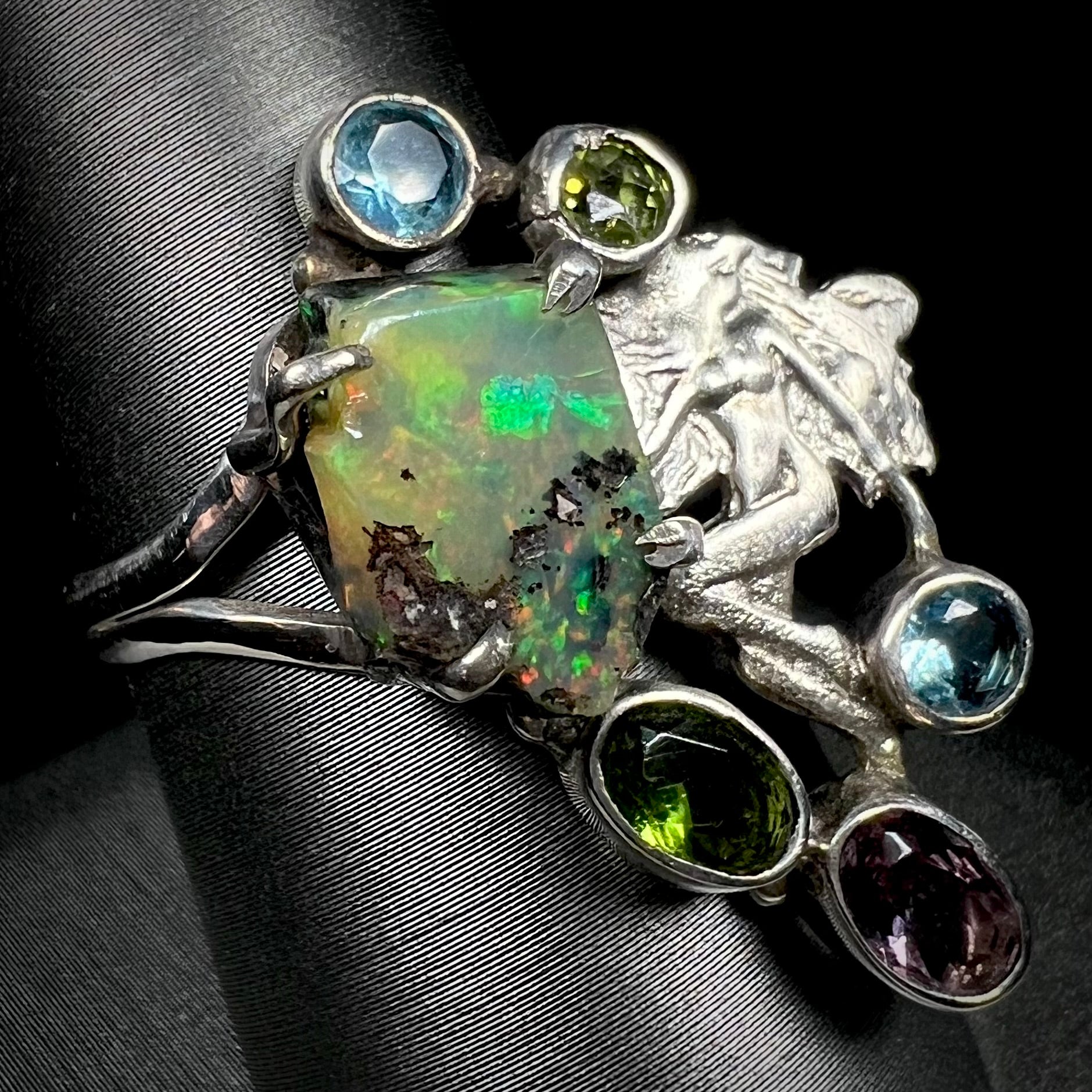 A silver men's ring set with rough Virgin Valley opal and gems with a female figure design shown from an angle.