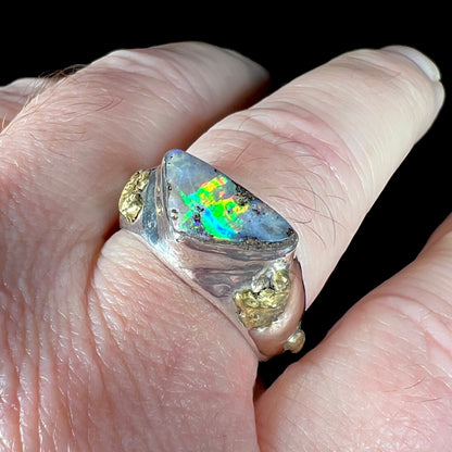 Angled close-up view of a men's size 12 sterling silver and gold nugget opal ring, worn on a man's finger.  The opal has bright green flashes of color in studio light.