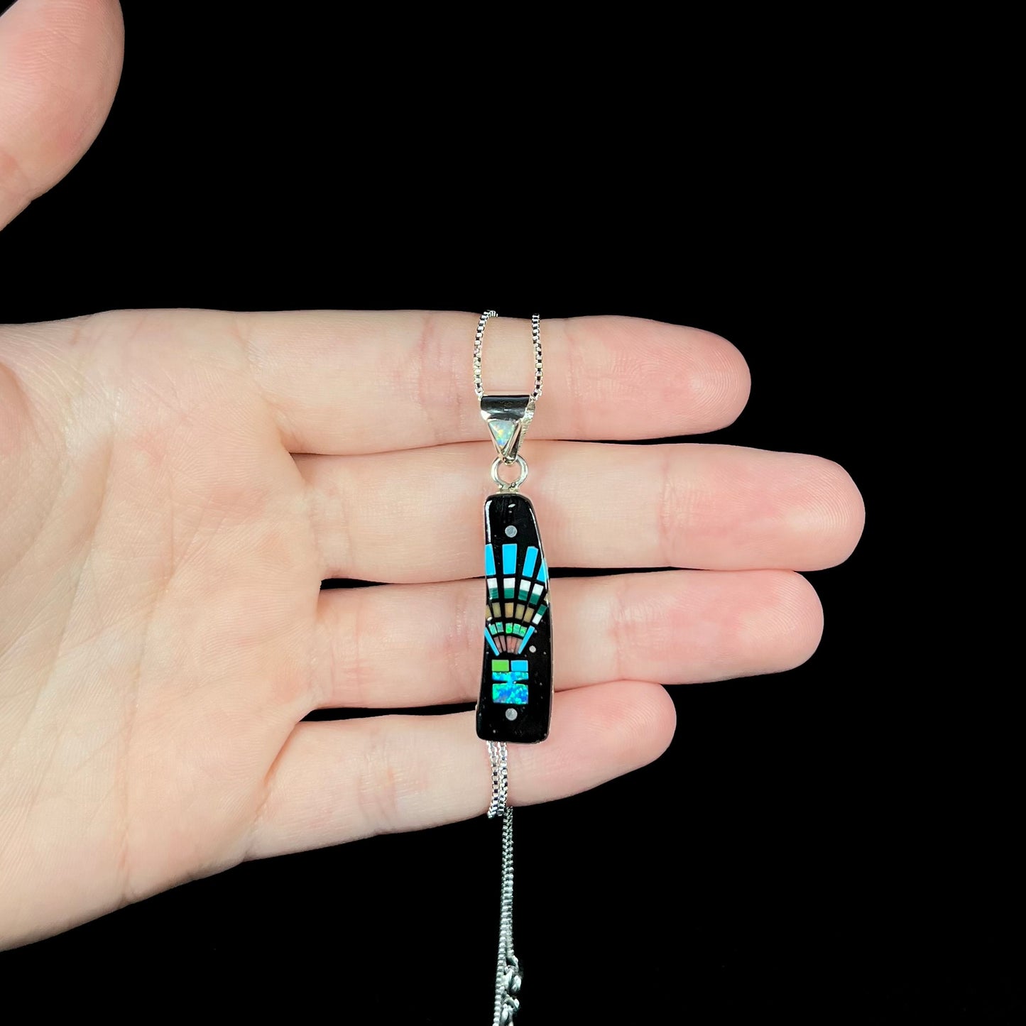 Hand holding a men's Southwest-style sterling silver opal in onyx inlay necklace against black background under cool LED studio lights.