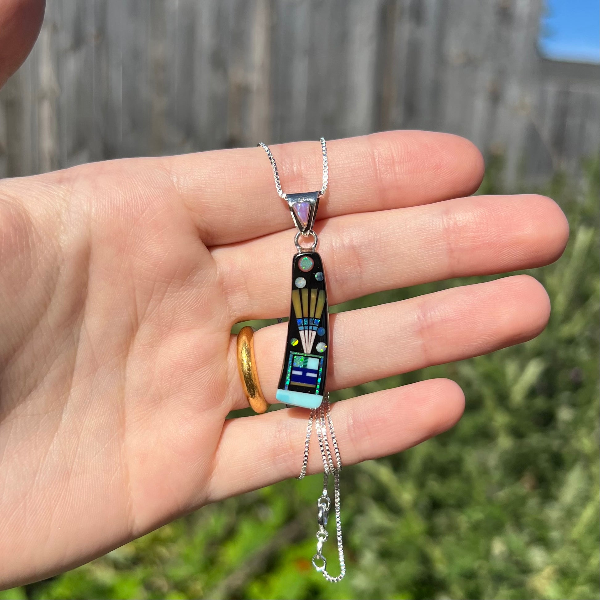 Hand holding men's southwest sterling silver opal and black onyx inlay necklace in natural sunlight against blurred outdoor background.