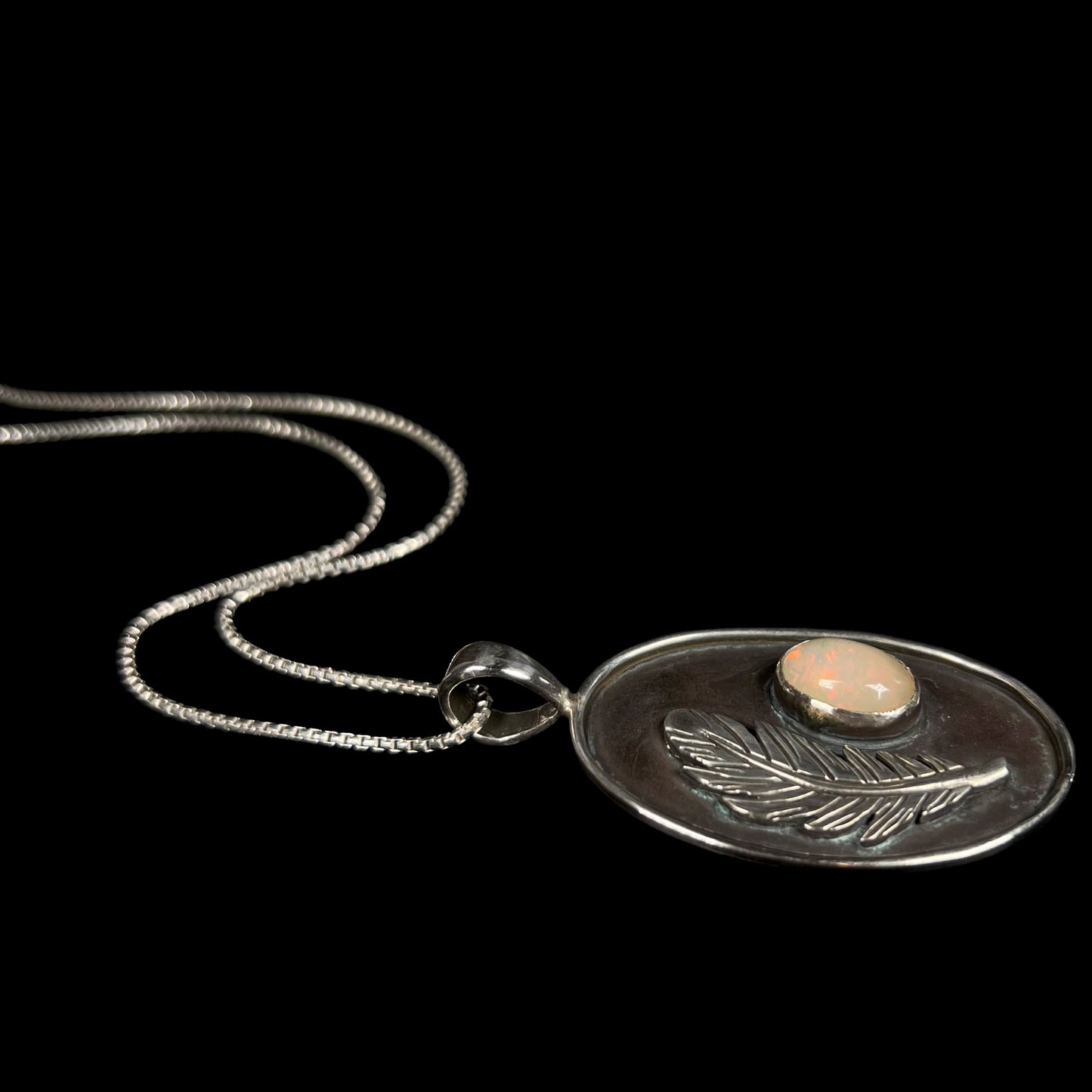 Angled view of a men's sterling silver Ethiopian opal and feather design necklace, laying against a black background.
