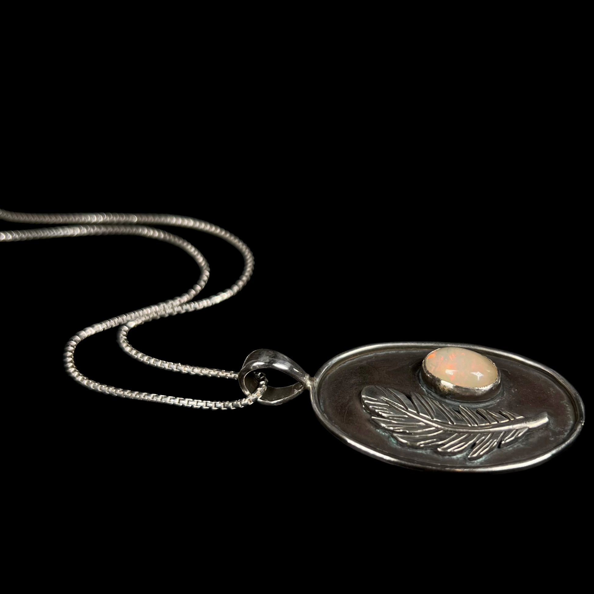 Angled view of a men's sterling silver Ethiopian opal and feather design necklace, laying against a black background.