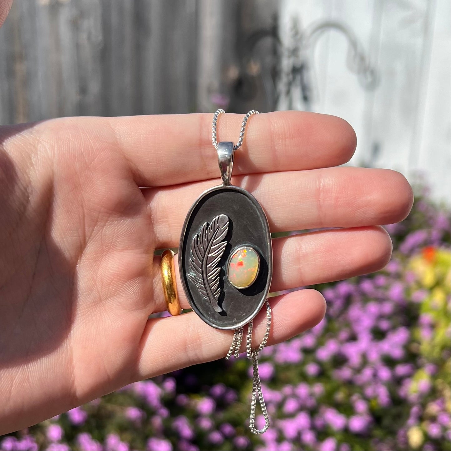 Hand holding a men's sterling silver fire opal and feather design necklace with red play of color in sunlight against blurred purple flower background.