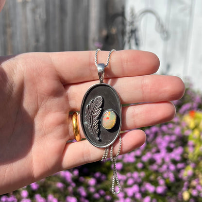Hand holding a men's sterling silver fire opal and feather design necklace with red play of color in sunlight against blurred purple flower background.