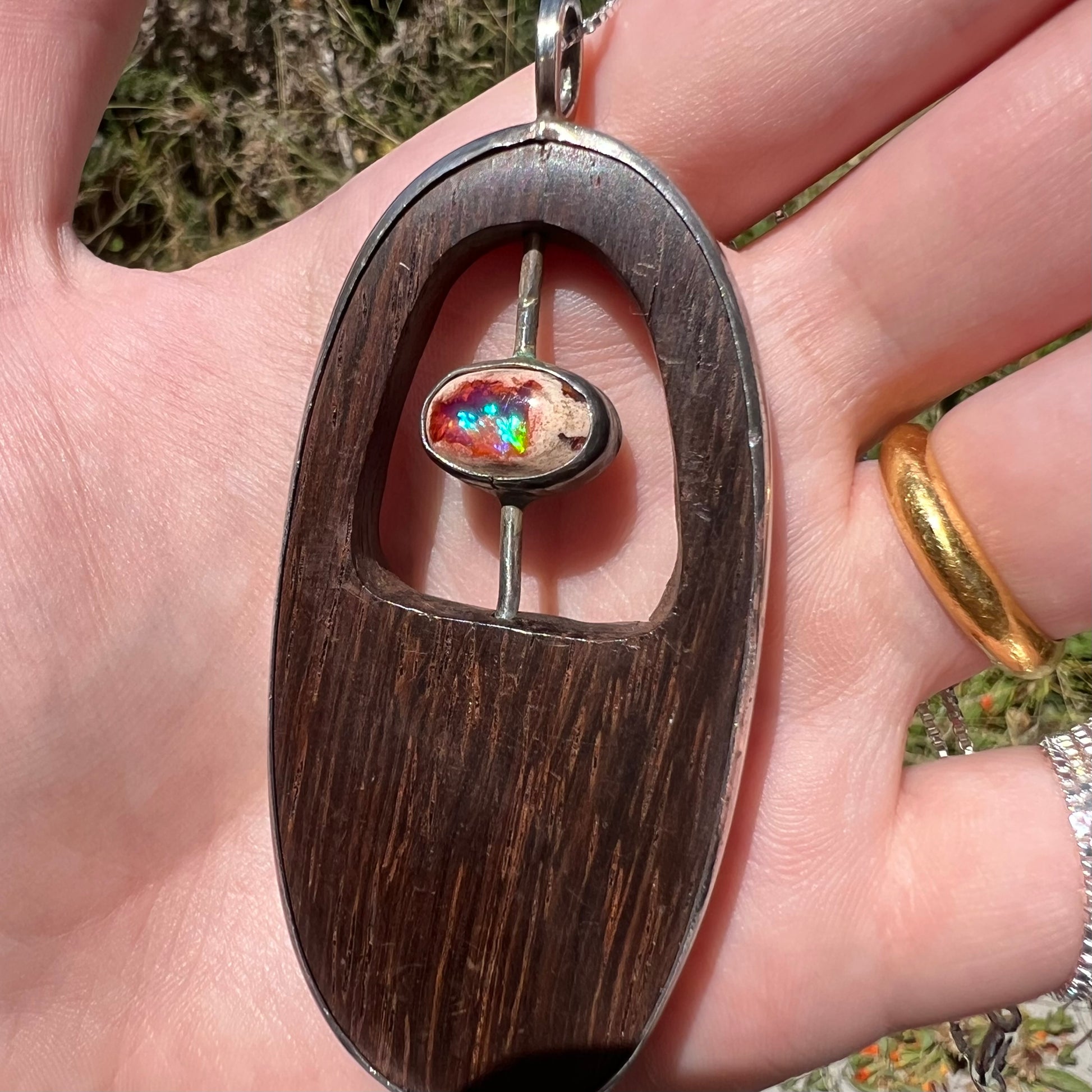 Angled close-up of a men's sterling silver fire opal and carved wood pendant, held in a hand in natural light.  The opal has a blue flash of color.