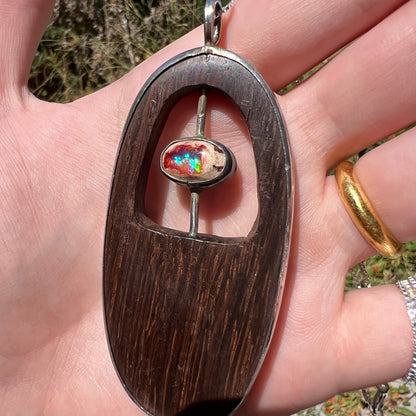 Angled close-up of a men's sterling silver fire opal and carved wood pendant, held in a hand in natural light.  The opal has a blue flash of color.