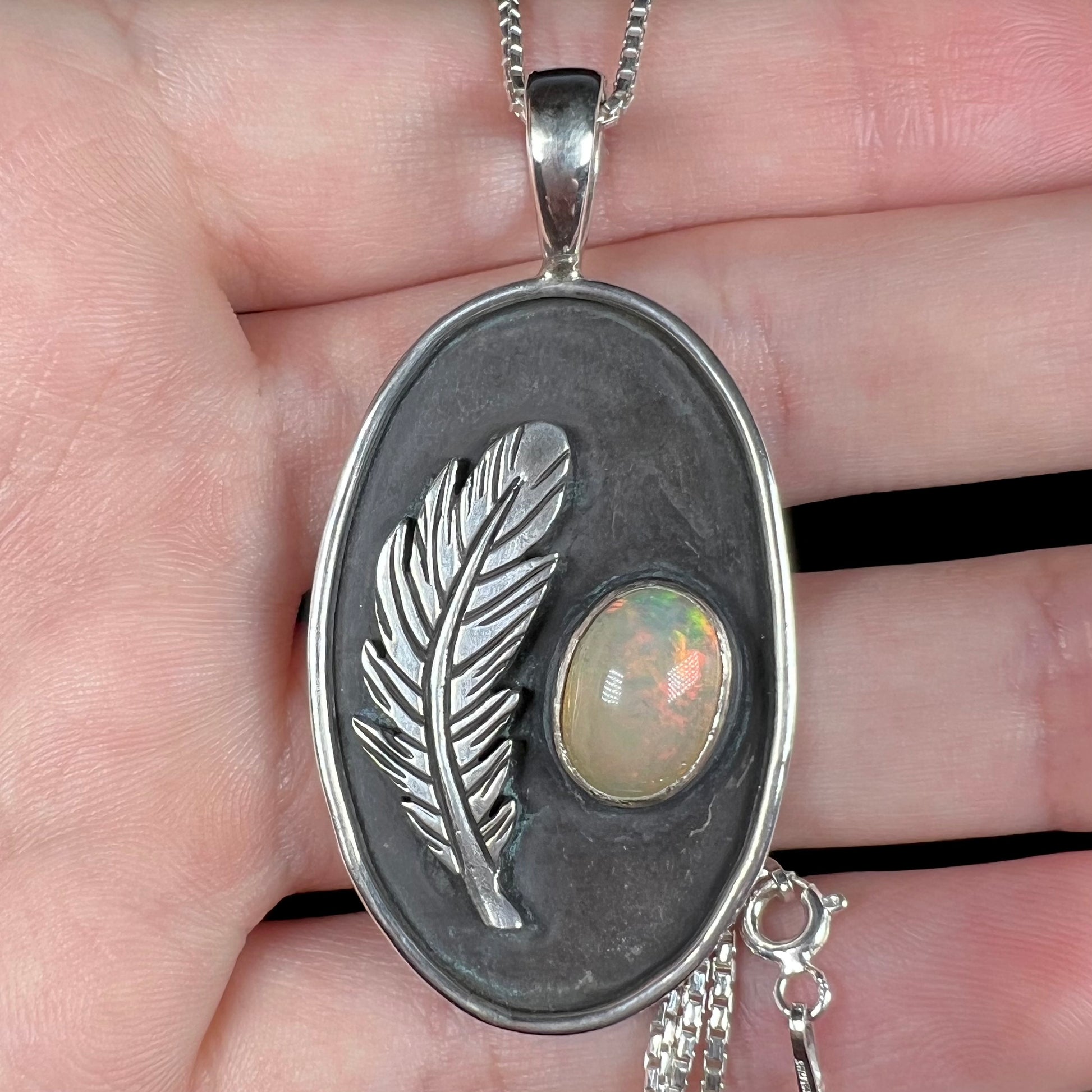 Close-up of a men's sterling silver opal and feather design pendant held in hand under cool LED studio light.