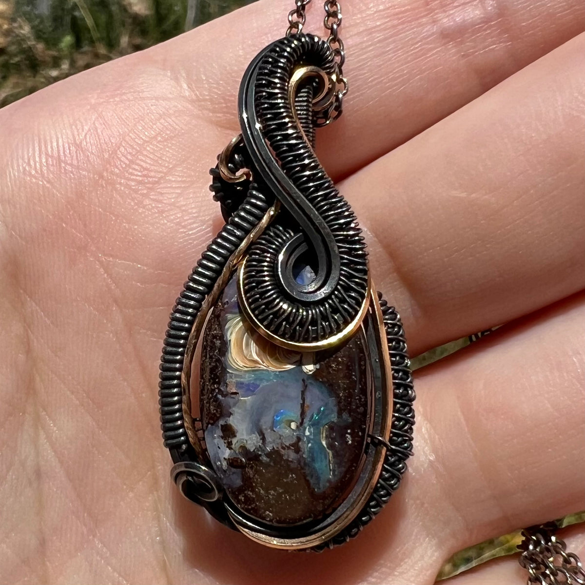 Close-up of a wire wrapped Australian boulder opal men's pendant in blackened sterling silver, shown in a hand under natural light.