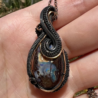 Close-up of a wire wrapped Australian boulder opal men's pendant in blackened sterling silver, shown in a hand under natural light.