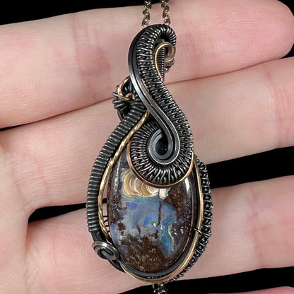 Close-up macro view of a men's wire wrapped Australian boulder opal pendant featuring two-tone black and gold-tone design, shown in a hand.