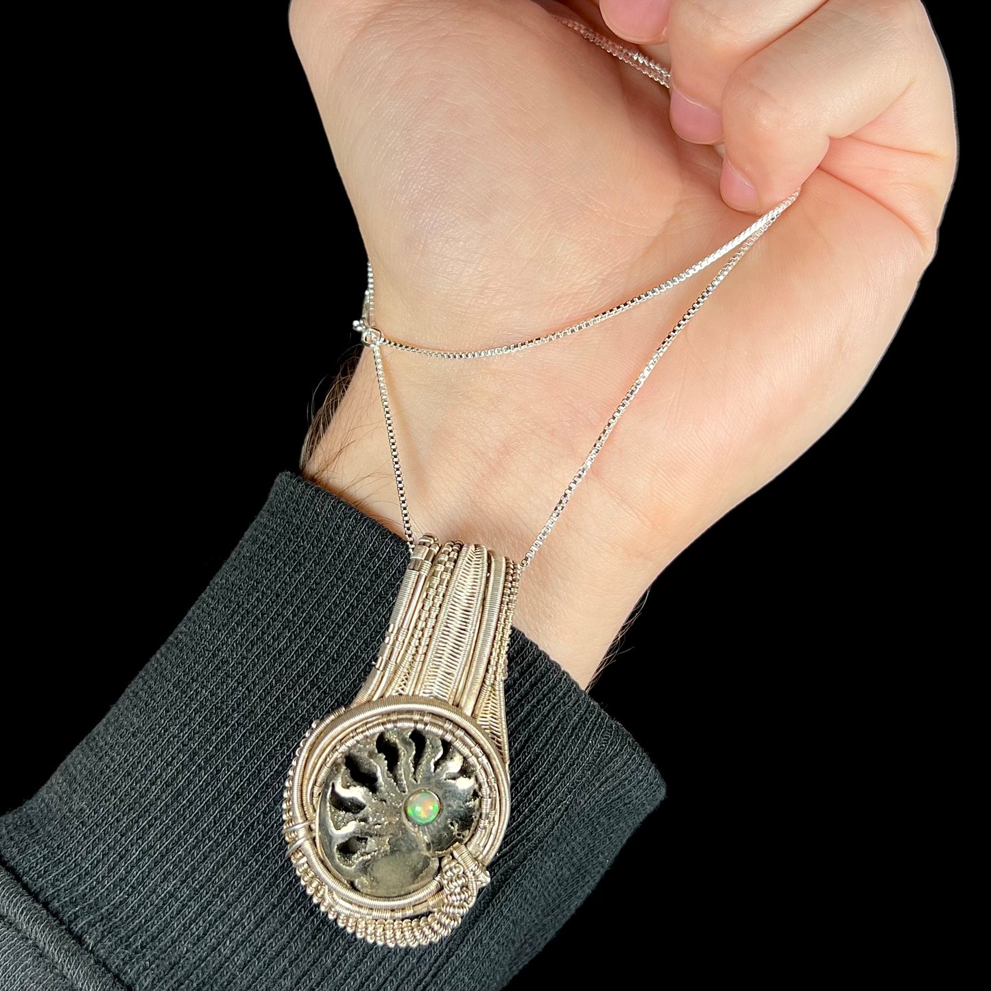 Hand holding men's wire wrapped opal-accented ammonite necklace from silver box chain so that pendant rests on wrist.