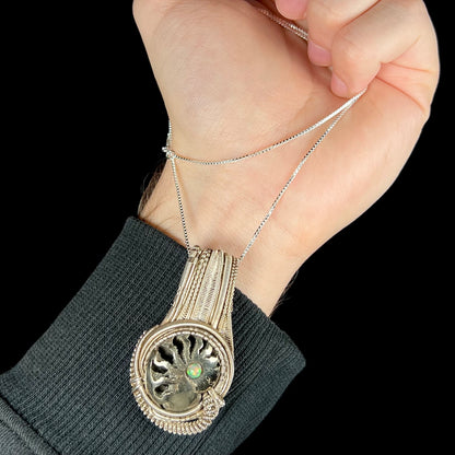 Hand holding men's wire wrapped opal-accented ammonite necklace from silver box chain so that pendant rests on wrist.