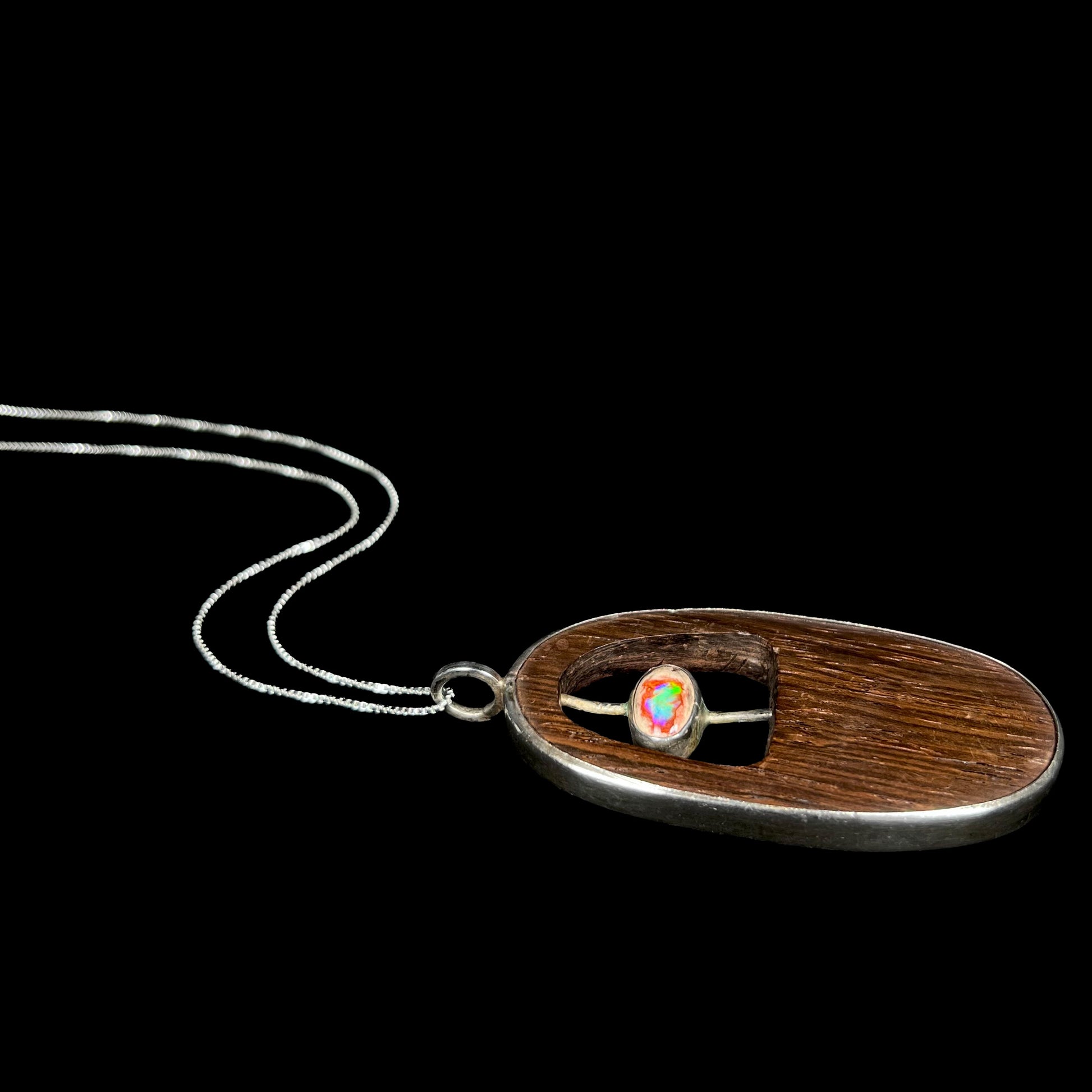 A sterling silver Mexican cantera fire opal and wood necklace for men shown from an angle laying on a black background.  The opal has a green flash.