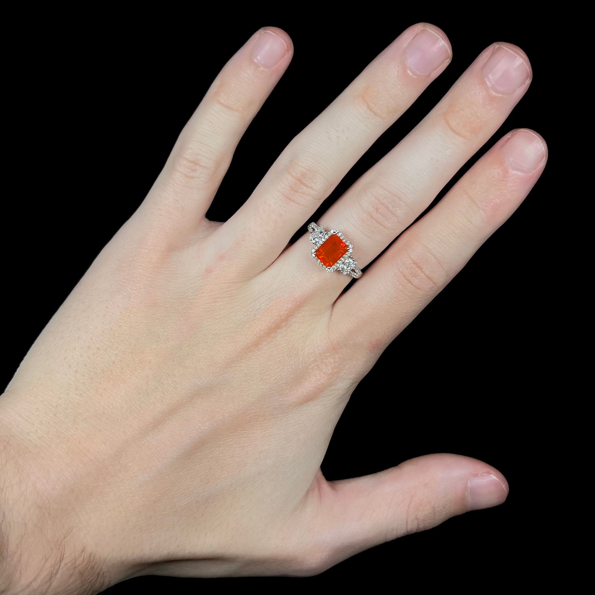 Hand wearing an orange Mexican fire opal and zircon halo ring under studio LED light against a black background.