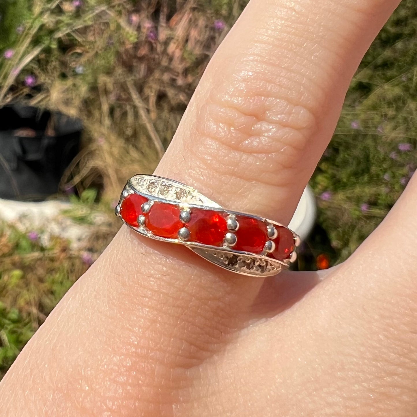 Macro front view of silver Mexican fire opal and diamond ring worn on a pinky finger in sunlight.