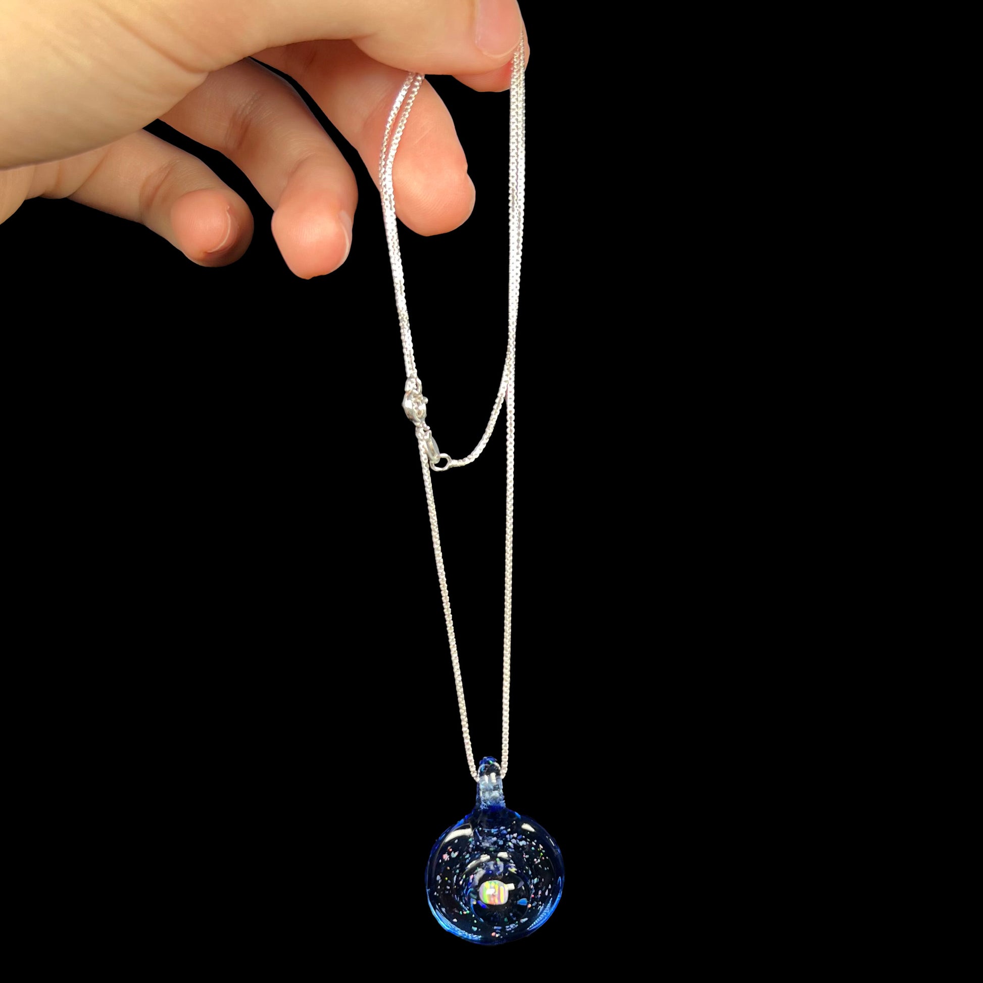 A Michigan-made glass and lab-created opal necklace, dangled by a hand from its silver chain against a black background.