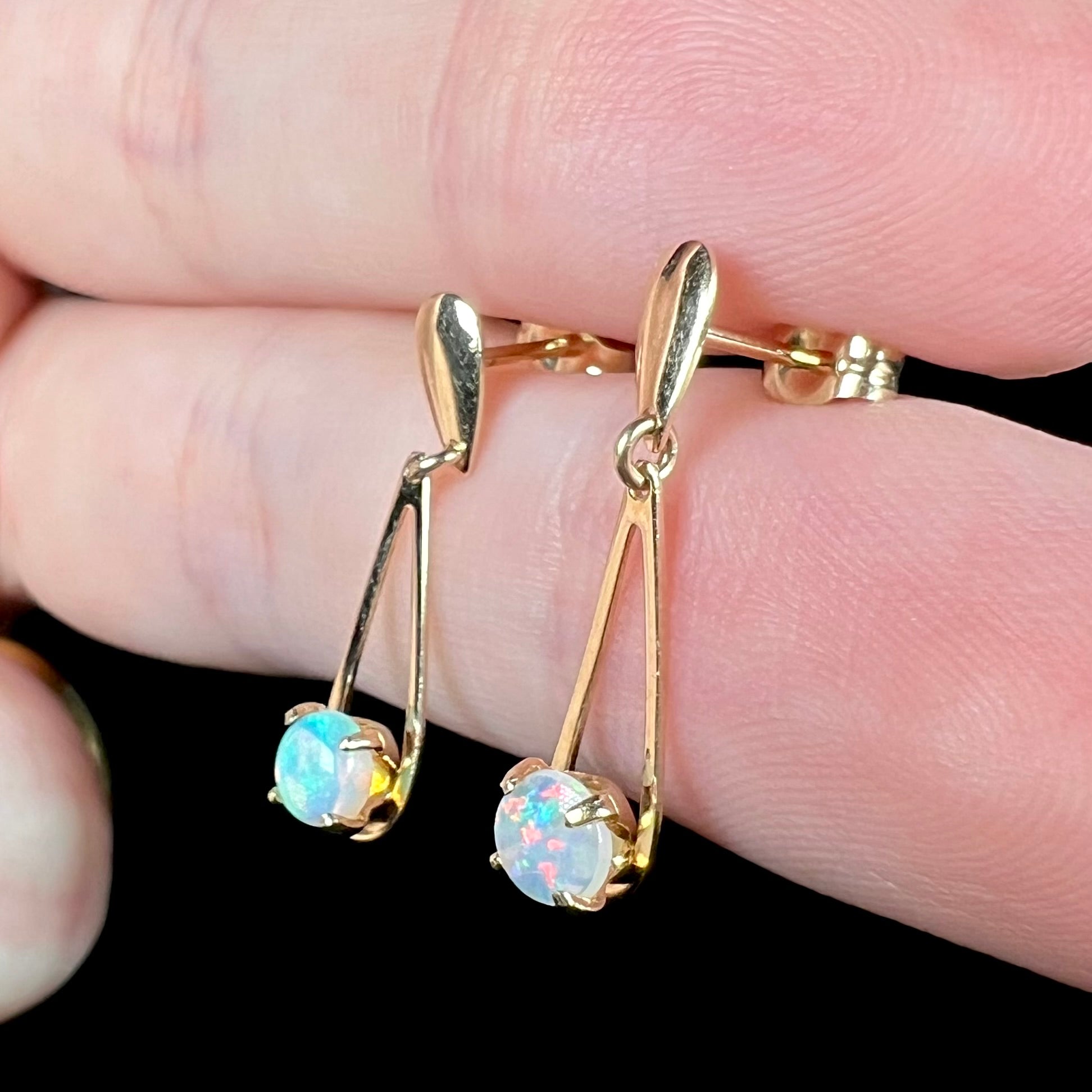 Pair of mismatched red and green Australian opal dangle earrings in gold held between two fingers from angle under cool studio lighting.