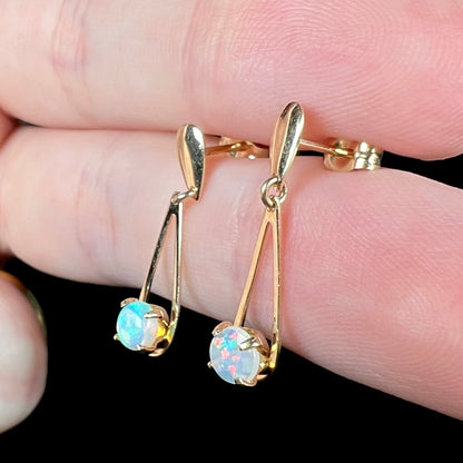 Pair of mismatched red and green Australian opal dangle earrings in gold held between two fingers from angle under cool studio lighting.