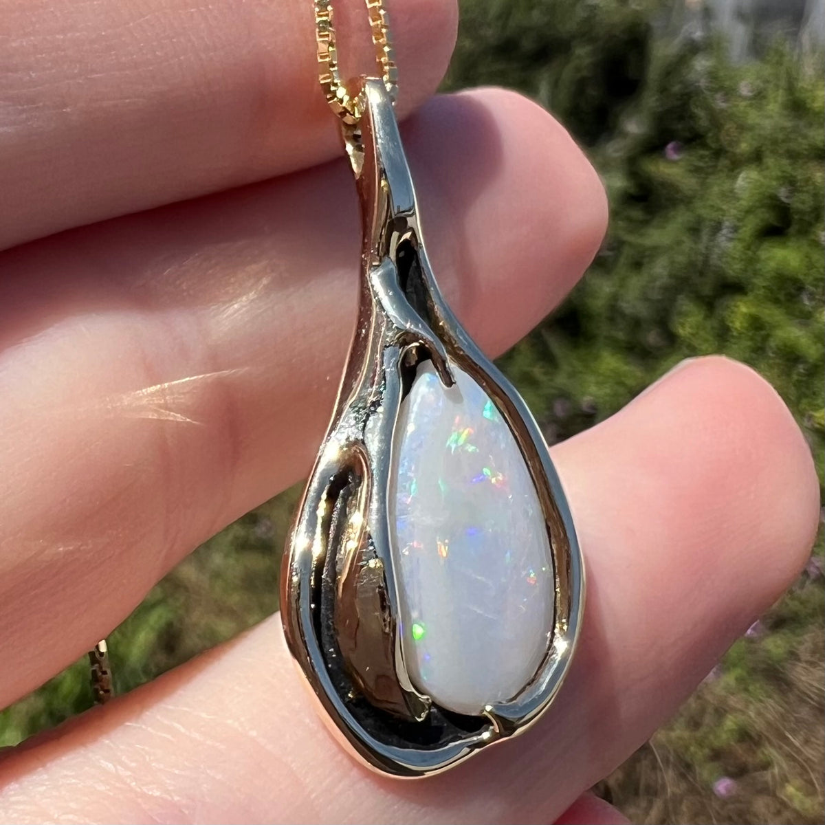 14k yellow gold Modernist style opal pendant shown in sunlight held in hand, the camera focused on the opal beside a section of black antiquing to its right