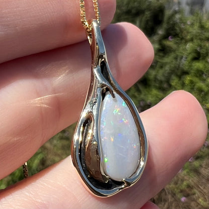 14k yellow gold Modernist style opal pendant shown in sunlight held in hand, the camera focused on the opal beside a section of black antiquing to its right
