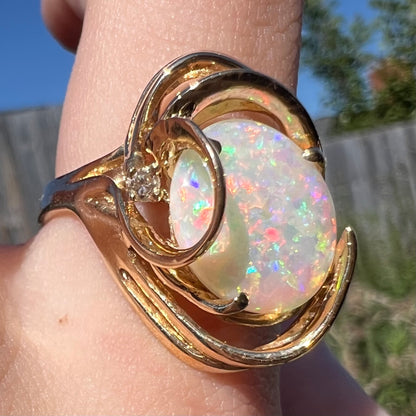 Angled macro view of Modernist style Australian opal and diamond cocktail ring with large, swooping 14k gold design, worn on finger in sunlight