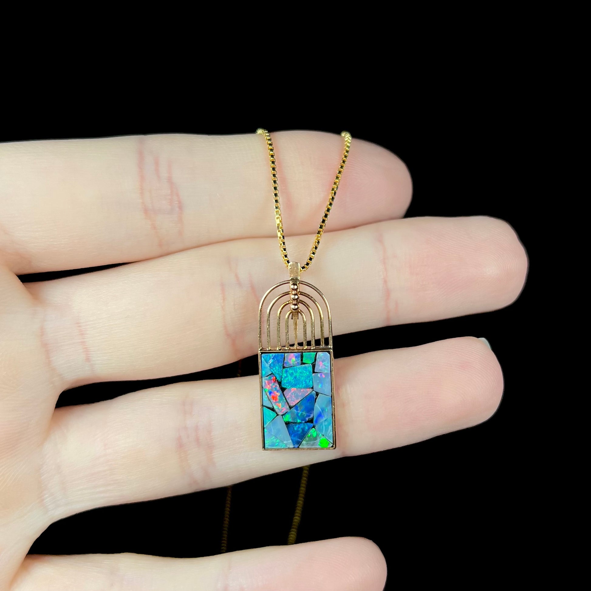 A mosaic opal pendant resting on fingers against a black background, with red and green flashes in the opal inlays.