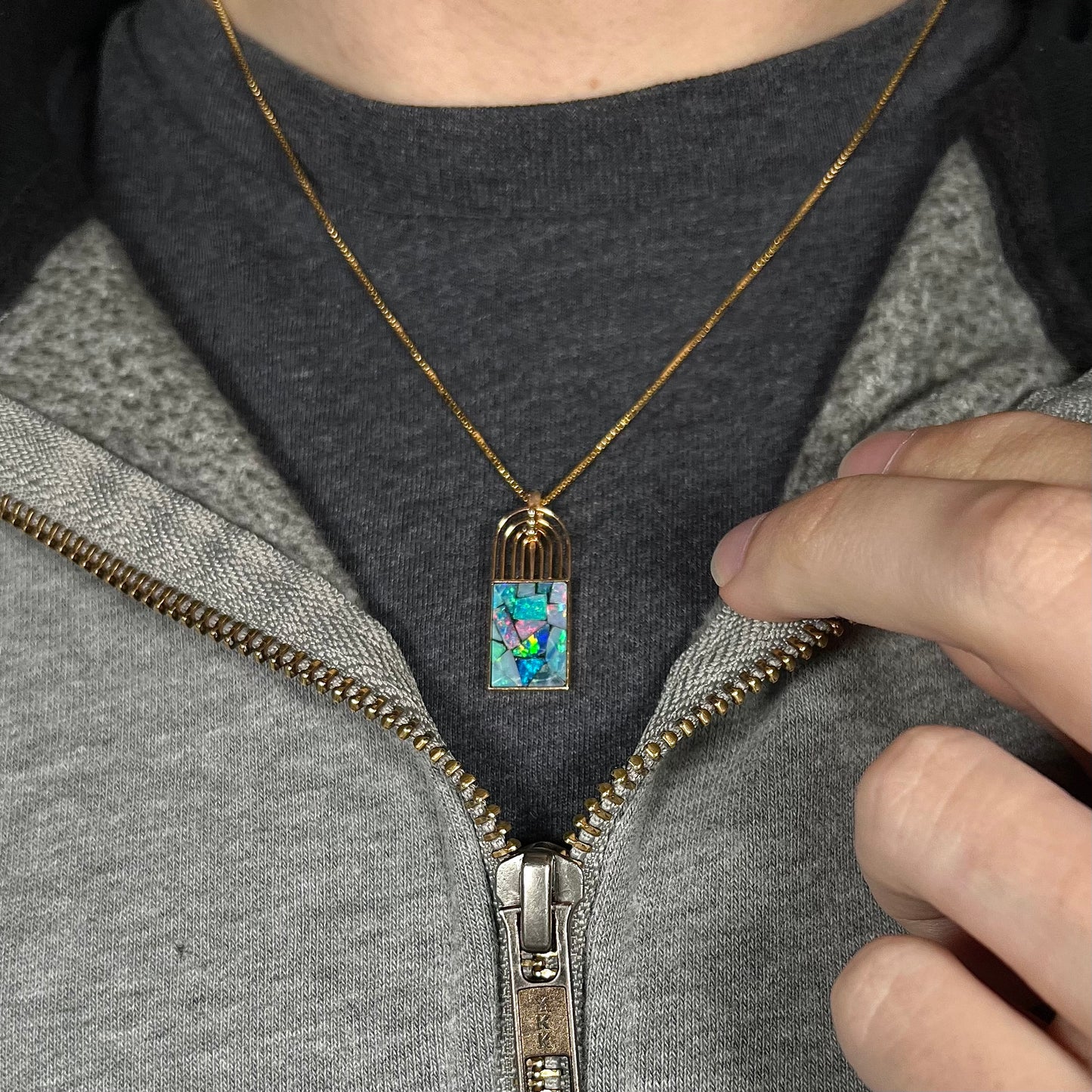 An 18k gold mosaic opal necklace worn on a black t-shirt with a partially zipped sweatshirt, displaying vibrant flashes of color.