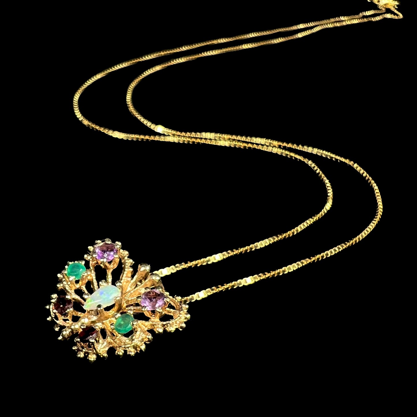 A multi-gemstone opal, amethyst, emerald, and garnet butterfly necklace in 14k gold shown from angle on black background with chain.